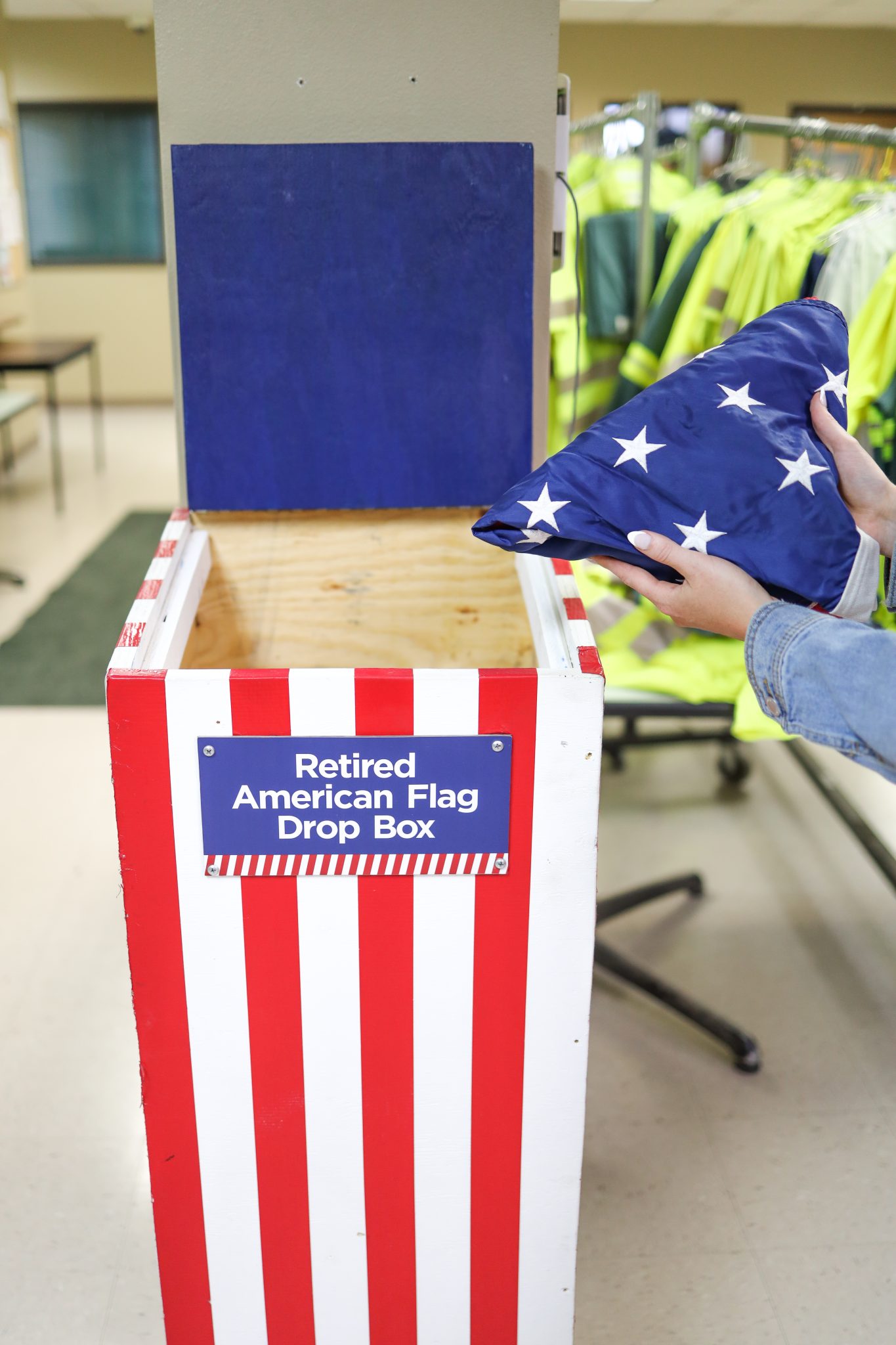 A red-white-and-blue box with the label, "Retired American Flag Drop Box" on it. A person is putting a flag in the box.