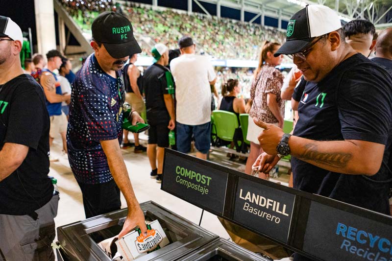 People inside Austin FC's Q2 Stadium disposing trash in front of three separate bins that read Compost, Landfill, and Recycle